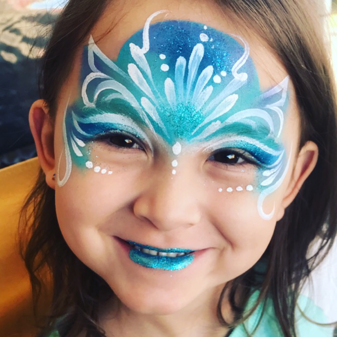 A blue and white whimsical face paint on a little girl with glittery blue lipstick