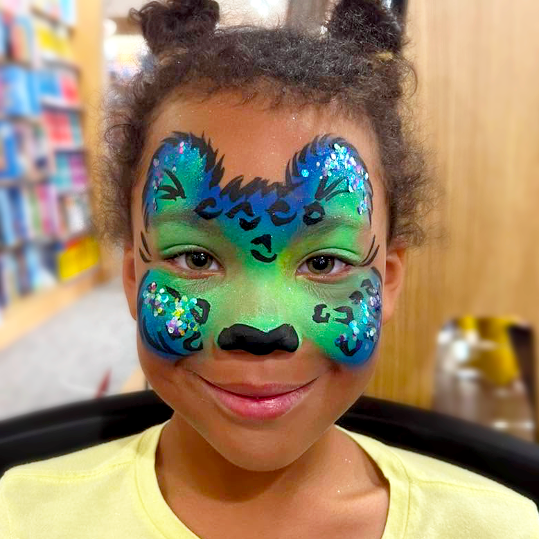A little girl with her face painted as a blue and green glittery leopard.